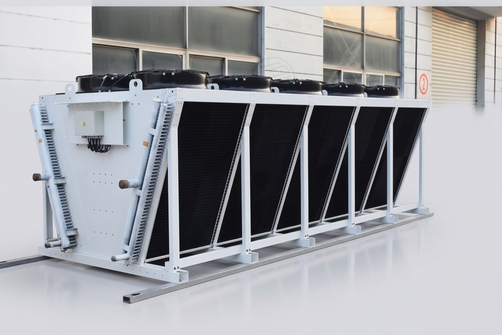 Air-cooled condenser with six axial fans and coated coil slabs in a V-bank on a steel frame, installed outside a plant building.